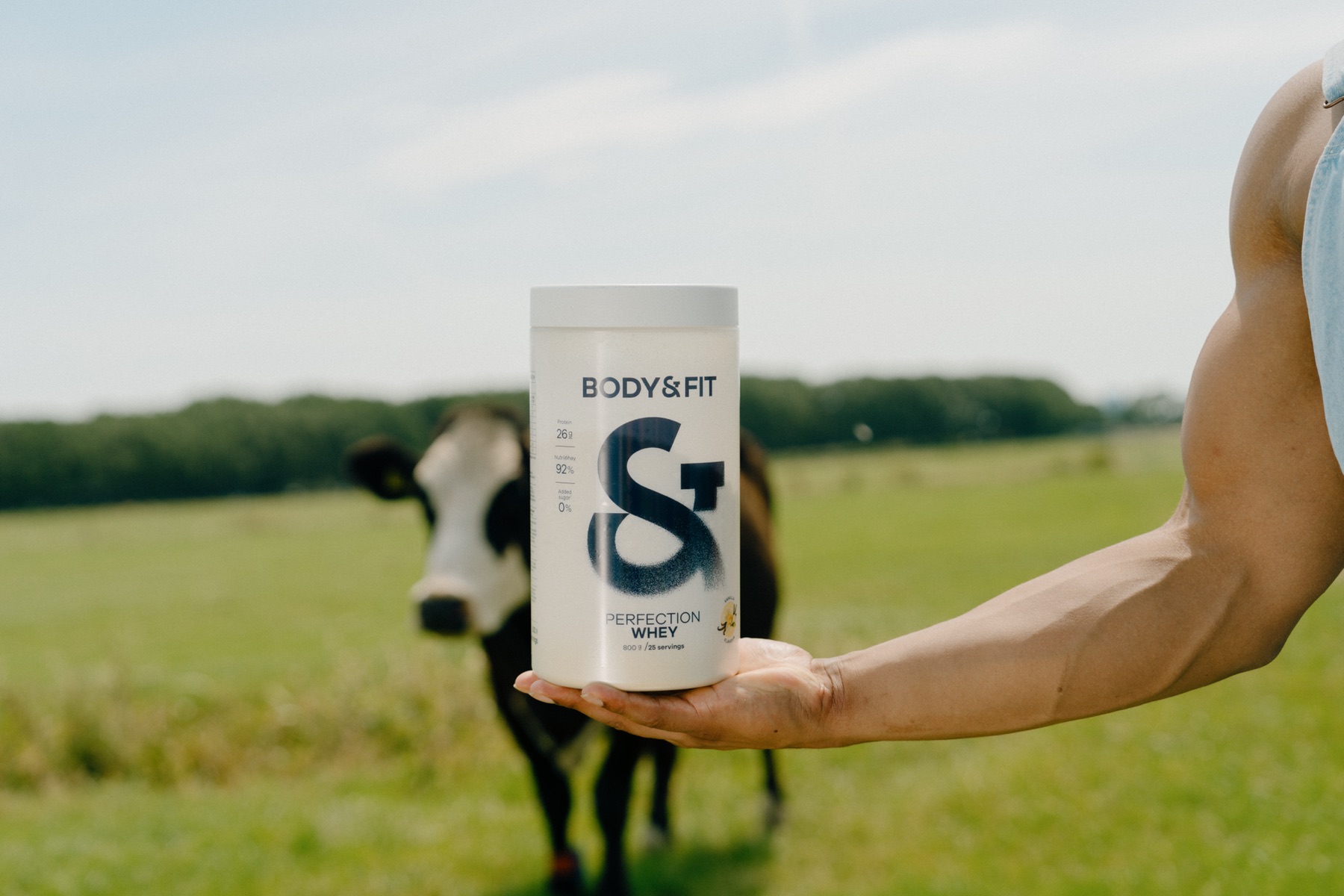 Hand houdt een Body&Fit Perfection Whey-bus vast met een koe in de wei op de achtergrond